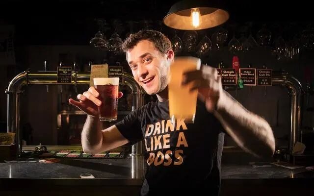 Dieter 在吧台后笑着举起两杯啤酒,T 恤上写着 DRINK LIKE A BOSS